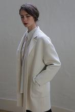 Load image into Gallery viewer, A person with short dark hair is standing against a plain, light-colored background. They are wearing a long, cream-colored Bamford Double-face Cashmere | Undyed coat over a white shirt. One hand is in a pocket of the coat, and they have a composed expression with their gaze directed downward.