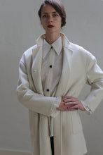 Load image into Gallery viewer, A woman with dark hair, styled in a side bun, stands against a plain background. She is wearing the Bamford Double-face Cashmere | Undyed, layered over a sheer white blouse with a buttoned collar and black buttons. Her hands are clasped at her waist, and she has a serious expression.