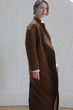 Load image into Gallery viewer, A person with fair skin and dark hair, styled in an updo, wears a Bamford Long Double-face Cashmere | Tobacco coat with a stand-up collar and large front pockets. They pose against a white textured wall, looking slightly past the camera with a poised expression.