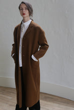 Load image into Gallery viewer, A person with short hair, wearing a Bamford Long Double-face Cashmere | Tobacco coat over a white button-up shirt and black pants, stands indoors against a light-colored wall. They have their hands in their coat pockets and are gazing directly ahead with a neutral expression.