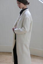 Load image into Gallery viewer, A person with tied-back hair is wearing a Bamford Long Double-face Cashmere | Undyed over a dark outfit. They are standing indoors against a neutral background, facing left, with hands held together at the waist. The setting has minimalistic decor with light wooden flooring and side patch pockets visible.