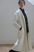 Load image into Gallery viewer, A person with long dark hair and red lipstick is wearing a Bamford Long Double-face Cashmere | Undyed coat with side patch pockets over a dark outfit. They are standing against a plain white wall, looking slightly to the left with one hand in their pocket.