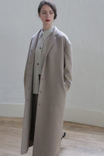 Load image into Gallery viewer, A person with dark hair tied back is wearing a long, light gray overcoat over a shirt and dark pants. They stand in a minimalistic indoor setting with a white wall and wooden floor, hands in pockets, looking off to the side, exuding an air of versatile elegance usually found in the Bamford Long | Undyed Natural.
