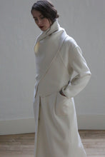 Load image into Gallery viewer, A person wearing a stylish, asymmetrical white coat with ceramic buttons and an X Scarf - Double-face Cashmere | Undyed stands against a plain wall with a wooden floor. The coat has large front pockets, and the individual has their hands in the pockets, looking down slightly. Their hair is tied back, and they have a calm expression.