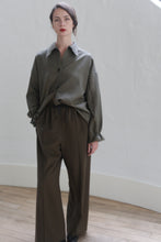 Load image into Gallery viewer, A person with long brown hair stands indoors against a white background. They are wearing an oversized grey button-up shirt with the sleeves rolled up and loose-fitting Cashmere Trousers | Olive. Accentuating their outfit, they have silk ribbon ties on their wrists. Their calm expression complements the relaxed pose of their hands by their sides.
