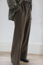 Load image into Gallery viewer, The image shows a person standing in Cashmere Trousers | Olive, paired with a loose, long-sleeved olive green top. The floor is wooden and the background features a white wall. The person is partially cropped out of the frame.