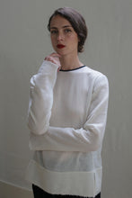 Load image into Gallery viewer, A woman with dark hair styled in an updo is standing against a plain background. She is wearing a Crewneck shirt - Japanese Wool Gauze | Undyed, reflecting sustainable design. Her left arm is crossed, with her right hand touching her chin as she looks straight at the camera with a serious expression.
