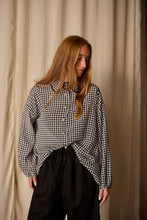 Load image into Gallery viewer, A woman with long, light brown hair is standing against a beige curtain backdrop. She is wearing a black and white houndstooth patterned Painter's Shirt | Crepe de Chine Houndstooth made of 100% silk crepe de chine and black pants. Her gaze is directed downwards and to the left.