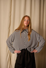 Load image into Gallery viewer, A woman with long blonde hair stands in front of a beige curtain. She is wearing Atelier Lan Jaenicke's Painter's Shirt | Crepe de Chine Houndstooth, crafted from 100% silk crepe de chine, tucked into black pants. Her hands are resting on her hips.