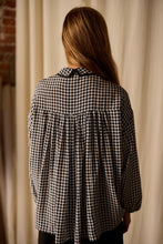 Load image into Gallery viewer, A person with long, light brown hair is standing with their back to the camera, wearing a black and white houndstooth patterned Painter's Shirt | Crepe de Chine Houndstooth with a pleated back. The blouse, 100% silk crepe de chine from Atelier Lan Jaenicke, contrasts beautifully against the light-colored curtain and partial brick wall.