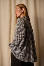 Load image into Gallery viewer, A person with long blond hair is captured in profile, wearing a loose-fitting, Painter's Shirt | Crepe de Chine Houndstooth. They are facing to the left in front of a plain, light-colored curtain background. The elegant top is crafted from 100% silk crepe de chine by Atelier Lan Jaenicke.