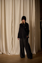 Load image into Gallery viewer, A person stands in front of beige curtains, wearing a loose-fitting Qipao Jacket | Cashmere Black with gold buttons and dark wide-legged pants. This elegant ensemble by Atelier Lan Jaenicke features a handmade touch. Their hands are in their pockets, and their face is partially obscured by the hood's shadow.