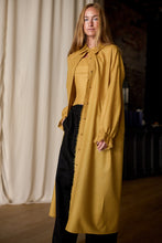 Load image into Gallery viewer, A person with long hair is wearing a Dress Coat | Japanese Wool Gauze Gold, over a beige top and black pants. They are standing indoors, framed by beige curtains in the background.