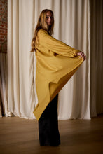 Load image into Gallery viewer, A person with long hair stands indoors, wearing a Dress Coat | Japanese Wool Gauze Gold that fans out as they hold it away from their body, revealing black pants underneath. The background features off-white draped curtains and a section of exposed brick wall.