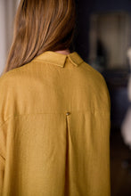 Load image into Gallery viewer, A person with long, light brown hair is seen from behind, wearing a Dress Coat | Japanese Wool Gauze Gold. The focus is on the back of the coat, which features a pleat secured by a small metal button. The background is softly blurred, showing indistinct indoor elements.