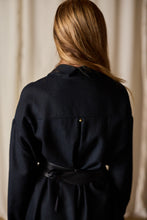 Load image into Gallery viewer, A person with long, light brown hair is pictured from behind, wearing a Dress Coat | Japanese Wool Gauze Black. The fabric appears to be soft and the top is cinched at the waist with a black ribbon or belt. The background is blurred and neutral in color.