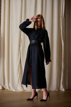Load image into Gallery viewer, A woman stands indoors in front of beige curtains, wearing a Dress Coat | Japanese Wool Gauze Black with a belt at the waist. She has one hand near her face and the other resting by her side. She is wearing black high heels and has straight, shoulder-length hair.