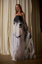 Load image into Gallery viewer, A person with long hair wears a Custom Shuimo Painting Dress | Satin Organza Buckhouse Print featuring an exclusive print in abstract black and white design. They stand on a wooden floor with cream-colored curtains in the background, their hand lightly grasping the custom-made fabric of the dress.
