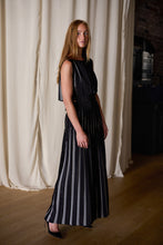 Load image into Gallery viewer, A woman with long red hair is standing in front of cream-colored curtains, wearing the Hand Pleated Column Dress Reversible | Custom Houndstooth Stripe and black heels. The 100% silk dress is handmade, giving it a unique touch. She has a relaxed expression and is looking slightly to the side against an indoor backdrop with wooden flooring.