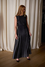 Load image into Gallery viewer, A woman with long, light brown hair stands indoors in front of off-white curtains. She is wearing a sleeveless, floor-length Hand Pleated Column Dress Reversible | Custom Houndstooth Stripe and black high heels. She looks to the side with one hand holding a part of her dress.