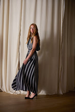 Load image into Gallery viewer, A woman with long hair wearing a Hand Pleated Column Dress Reversible | Custom Houndstooth Stripe stands against a backdrop of tall, white curtains. The dress, 100% silk and handmade, flows out gracefully as she faces slightly to the side, looking back at the camera.