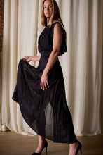 Load image into Gallery viewer, A woman with long red hair stands indoors against a curtain backdrop. She is wearing a handmade, black, sleeveless, floor-length Hand Pleated Column Dress | Black Cotton Silk Voile and black high heels. She holds one side of her dress up slightly, showcasing its flowing design.