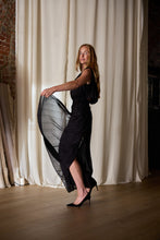 Load image into Gallery viewer, A woman with long red hair twirls in a flowing, sleeveless black Hand Pleated Column Dress crafted from cotton-silk voile and dons black high heels. She stands on a wooden floor in front of white curtains and a brick wall, with fabric rolls leaning against the wall in the background.