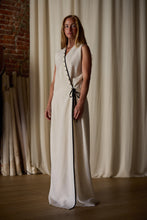 Load image into Gallery viewer, A person with long, light brown hair stands in a room with a brick wall and cream-colored curtains. They are wearing the Wrap Dress | Japanese Wool Gauze Undyed. The lighting is warm and soft, accentuating the serene ambiance inspired by sustainable design.