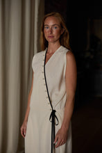 Load image into Gallery viewer, A woman with long auburn hair is standing indoors in a well-lit room with dark surroundings. She is wearing a sleeveless, light-colored Wrap Dress | Japanese Wool Gauze Undyed, featuring black silk trim and button details along the front. She has a calm, composed expression and is gazing into the camera.