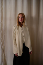 Load image into Gallery viewer, A woman with long blonde hair stands in front of beige curtains. She is wearing a sophisticated cream-colored Qipao Jacket | Cashmere Undyed, featuring an undyed silk lining and buttoned at the top while draping open at the bottom, paired with a dark skirt. The Qipao Jacket | Cashmere Undyed is handmade in-house, and she has a composed expression on her face.
