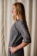 Load image into Gallery viewer, A woman with long, straight red hair is standing in profile against a light curtain backdrop. She is wearing a Qipao Dress | Japanese Wool Gauze Houndstooth with black trim along the seams and short sleeves. Her right hand is resting on her hip.