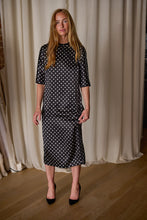 Load image into Gallery viewer, A woman stands on a wooden floor against a backdrop of light-colored curtains. She wears a black Polkadot Silk Charmeuse Qipao Dress with white polka dots that falls below her knees, complemented by black pointed-toe shoes. Her long, light brown hair cascades gracefully, and she exudes a calm expression.