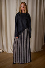 Load image into Gallery viewer, A person stands in front of draped curtains, wearing a Crewneck shirt - Japanese Wool Gauze | Black and a floor-length gray skirt with vertical black stripes. Crafted from Japanese wool gauze, this handmade ensemble complements their long blonde hair and neutral expression against the light-colored background and wooden floor.