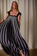 Load image into Gallery viewer, A woman stands in front of light-colored curtains, wearing a flowing black Hand Pleated Peony Dress Reversible | Custom Houndstooth Stripe with white vertical stripes and intricate patterns. The dress, crafted from luxurious silk Crepe de Chine, features a square neckline and cap sleeves. She gazes towards the camera with a gentle expression as the dress billows around her.
