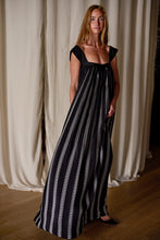 Load image into Gallery viewer, A woman with long hair stands indoors in front of white curtains. She is wearing a flowy, sleeveless black Hand Pleated Peony Dress Reversible | Custom Houndstooth Stripe adorned with white vertical stripes and a wide square neckline. Her hands rest in the dress's pockets, and she is looking confidently at the camera.