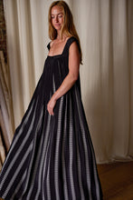 Load image into Gallery viewer, A woman with long hair, wearing a flowing Hand Pleated Peony Dress Reversible | Custom Houndstooth Stripe in black with vertical white stripes, stands in front of a beige curtain backdrop. She has a calm expression and her head is slightly tilted to one side. The room has a warm, wooden floor.