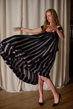Load image into Gallery viewer, A woman with long hair gracefully dances, swirling the skirt of her black and white striped off-the-shoulder Hand Pleated Peony Dress Reversible | Custom Houndstooth Stripe. She is wearing black high heels and stands in front of white curtains on a wooden floor.