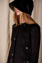 Load image into Gallery viewer, A woman with long hair stands against a neutral background, wearing the Crop Cashmere Jacket | Black with gold buttons and a large black hat that shades her eyes. Handmade in San Francisco, the jacket features two front pockets, and the overall look is minimalist and stylish.