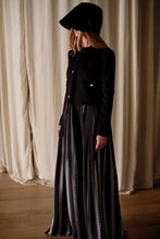 Load image into Gallery viewer, A person is standing indoors, dressed in a long, black and white vertical striped skirt, paired with a handmade Crop Cashmere Jacket | Black adorned with gold buttons. Their head is covered with a black hat casting a shadow over their face. The background features light, pleated curtains.