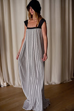 Load image into Gallery viewer, A woman stands wearing a black sleeveless top and a long, flowing silk crepe de chine skirt with a black and white zigzag pattern. She also has a black hat partially covering her head. The background features light-colored curtains and a hardwood floor, perfectly complementing her elegant Hand Pleated Peony Dress | Crepe de Chine Custom Houndstooth-inspired look.