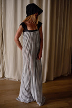 Load image into Gallery viewer, A person stands indoors against floor-length white curtains, elegantly clad in a loose, floor-length Hand Pleated Peony Dress | Crepe de Chine Custom Houndstooth made of silk crepe de chine with a black and white dotted pattern. Their face is partially obscured by a dark hood attached to the dress. The setting has wooden flooring.