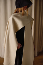 Load image into Gallery viewer, Person wearing a stylish, cream-colored Doubleface Cashmere X-Cape over a black outfit with a black wide-brimmed hat covering their face. The background features neutral, draped curtains.