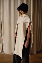 Load image into Gallery viewer, A person wearing a black wide-brimmed hat and a stylish off-white cape stands against a backdrop of beige curtains. The Doubleface Cashmere X-Cape drapes elegantly over their shoulders, handmade in-house for quality, offering multiple ways of styling as they gaze downward.
