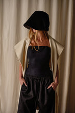 Load image into Gallery viewer, A woman stands against a neutral-colored backdrop. She is wearing a black, wide-brimmed hat that partially obscures her face, a strapless black top, loose black pants, and a Doubleface Cashmere X-Cape draped over her shoulders. Her hands are in her pockets.