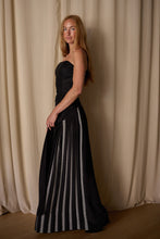 Load image into Gallery viewer, A woman with long hair is wearing an elegant, strapless black Layered Skirt w/ Hand Pleats | Crepe de Chine adorned with vertical white detailing on the skirt. The custom houndstooth stripe adds a unique touch. She is standing sideways, facing slightly towards the camera, with a neutral expression. The background features soft, cream-colored drapes.