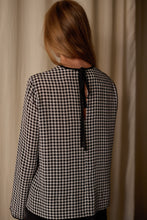 Load image into Gallery viewer, A person with long, light brown hair is standing facing away from the camera. They are wearing a black and white houndstooth patterned Qipao Blouse | Crepe de Chine Houndstooth, featuring a black ribbon tie at the neck. The background is a cream-colored curtain.
