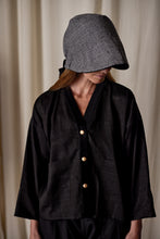 Load image into Gallery viewer, A person is wearing a dark, loose-fitting jacket with three light-colored buttons down the front and large pockets. They have on a Cashmere Ribbon Cloche | Black, Reversible with a wide brim covering their face. The background is blurred with soft, light-colored curtains.