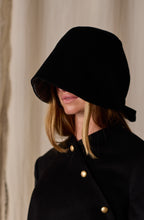 Load image into Gallery viewer, A person with long, blonde hair wears a Cashmere Ribbon Cloche | Black, Reversible that obscures their face. They are dressed in a black outfit featuring gold buttons. The background is a neutral, light-colored curtain.