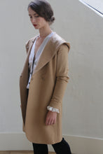 Load image into Gallery viewer, A person stands indoors next to a white wall, wearing a Mitchley Long | Camel with a hood. The person has dark hair pulled back and is looking down with a calm expression. They are wearing a white blouse underneath the coat, which features handmade snap buttons.