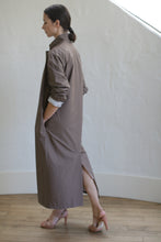 Load image into Gallery viewer, A person stands sideways on a wooden floor, wearing the Painter's Coat in Clay, cinched with a leather Obi belt and paired with pink heels. Their hair is tied back, one hand resting in a pocket. In the background is a white, curved wall softly illuminated by gentle lighting.